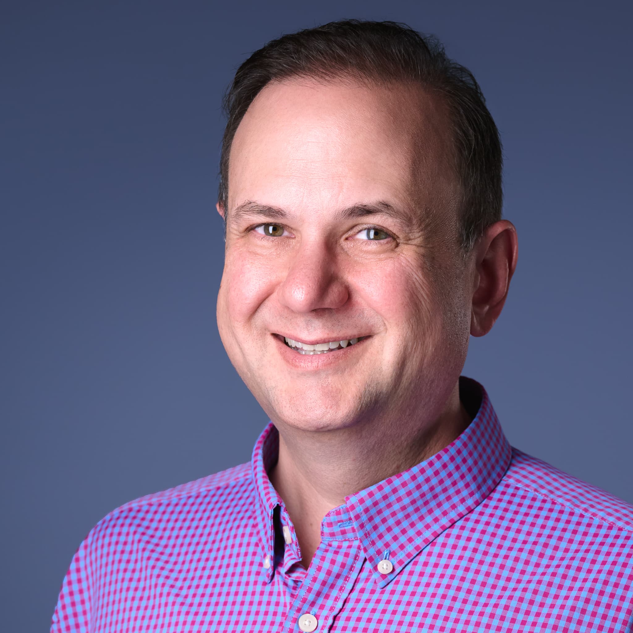 Color headshot in a pink and blue gingham shirt against a slate-blue backdrop.