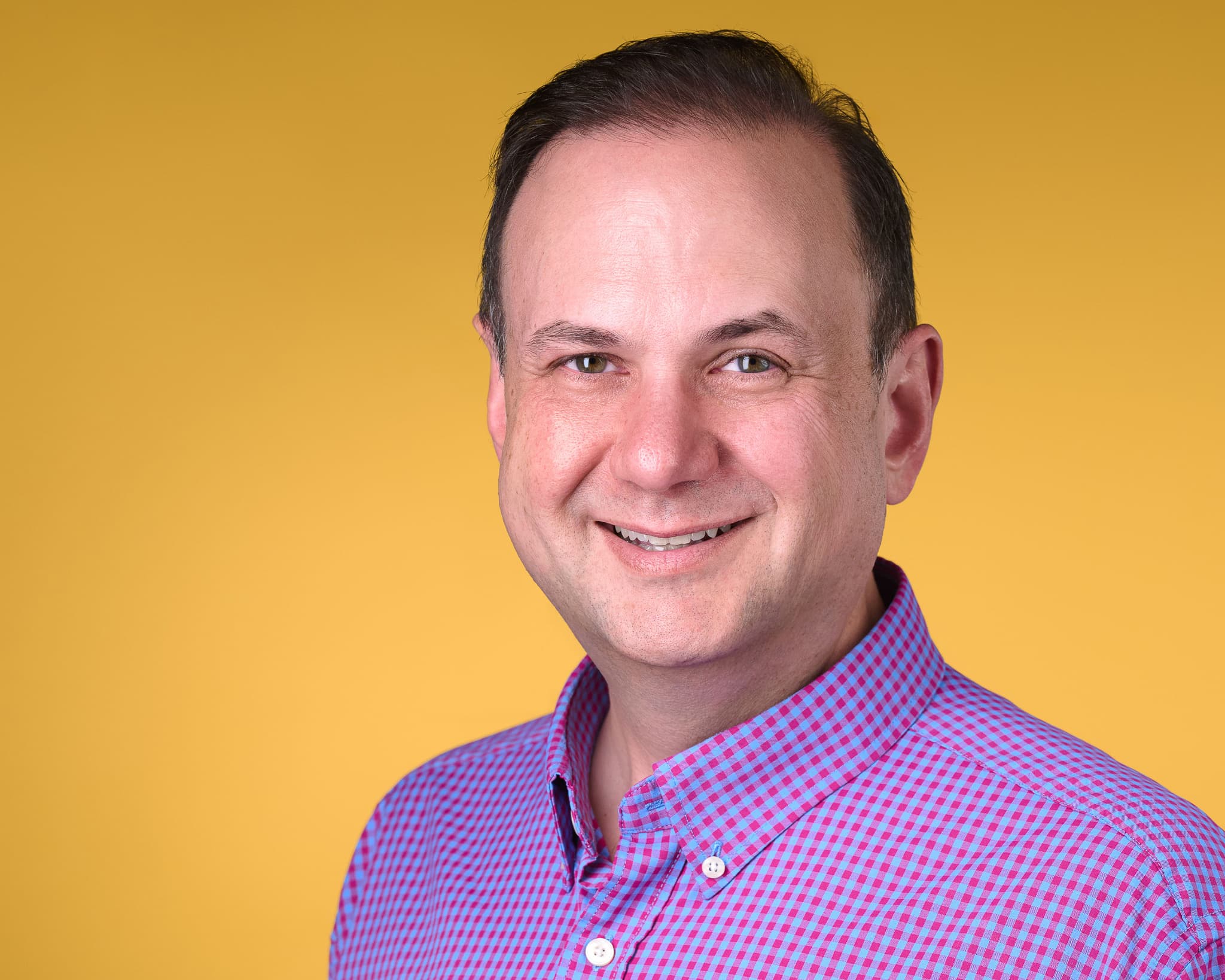 Color headshot in a pink and blue gingham shirt against a warm amber backdrop.